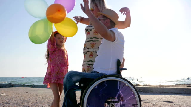 fröhliche-Mutter,-Tochter-mit-Luftballons-und-Papa-im-Gegenlicht,-kleine-Mädchen,-Mutter-und-Behinderten-Vater