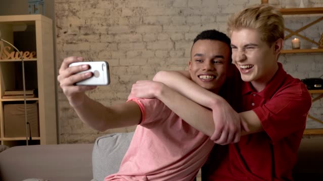 Two-international-gay-friends,-sitting-on-the-couch,-hugging,-making-selfies,-laughing.-Homeliness,-romantic-evening,-background,-hugs,-happy-LGBT-family-concept-60-fps