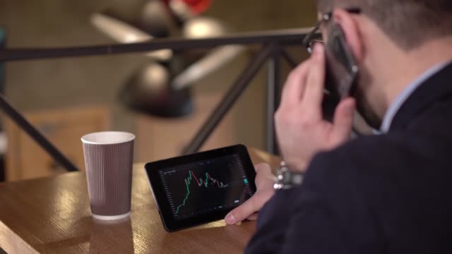 Junger-Mann-Monitore-Handel-Kryptowährung-auf-Tablet-und-Handy-in-einem-Café-spricht