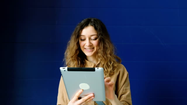 Mujer-joven-usa-una-tableta-para-comunicación-de-vídeo-sobre-un-fondo-de-pared-azul