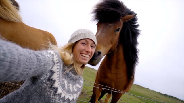 Blond-hair-girl-in-Iceland-taking-selfie-portrait-with-Icelandic-horse-in-green-meadow.-Shot-in-Springtime,-overcast-sky,-woman-wearing-Icelandic-grey-wool-pullover.-People-travel-animal-affection-concept--Slow-motion