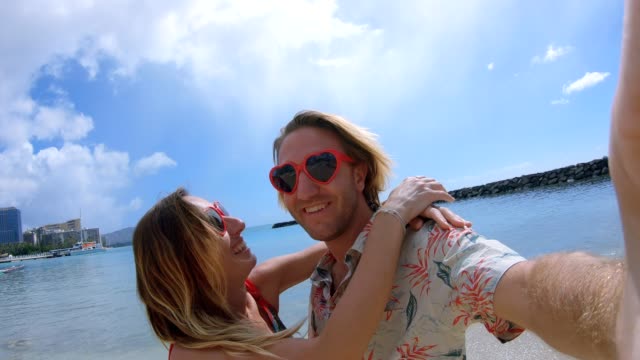 Par-tomar-autorretratos-en-la-playa-de-Waikiki-en-Honolulu,-Hawaii.-Selfie-granangular-punto-de-vista-de-la-playa-de-Waikiki.-Pareja-joven-tomar-autorretratos-con-el-corazón-en-forma-de-gafas-de-sol