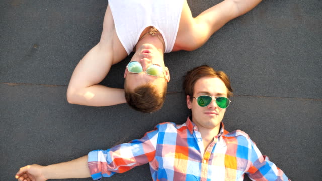 Top-view-of-young-happy-male-couple-in-sunglasses-lying-on-rooftop-of-high-rise-building-and-smiling.-Handsome-gay-boys-looking-at-camera-and-enjoying-to-spend-time-on-roof.-Close-up-Slow-motion
