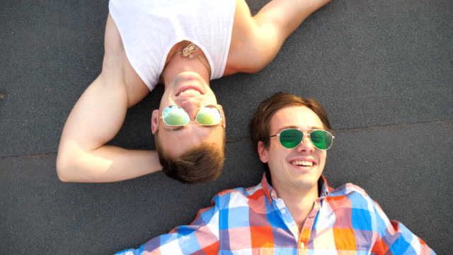 Junges-fröhliches-Männerpaar-in-Sonnenbrille-liegend-auf-dem-Dach-des-Hochhauses-und-freudiges-Lachen.-Hübsche-schwule-Jungs-schauen-sich-Kamera-an-und-lächeln,-während-sie-auf-dem-Dach-reden.-Blick-nach-oben