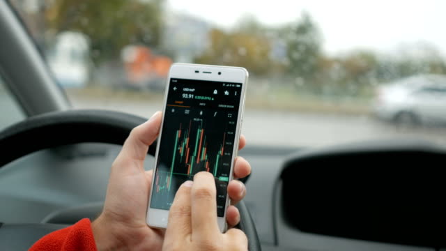 Driver-using-smartphone-device-to-check-market-data-in-the-car-on-city-background.-Stock-market,-trading-online,-trader-working-with-tablet-on-stockmarket-trading-floor.-Forex.-Crypto-currency.