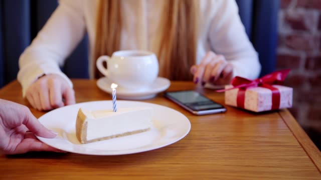 Close-up-of-waitress-serving-piece-of-birthday-cake-with-burning-candle-for-woman-having-tea-and-browsing-smartphone-alone-in-cafe,-waiting-for-somebody-to-come.-Birthday-gift-box-placed-on-table