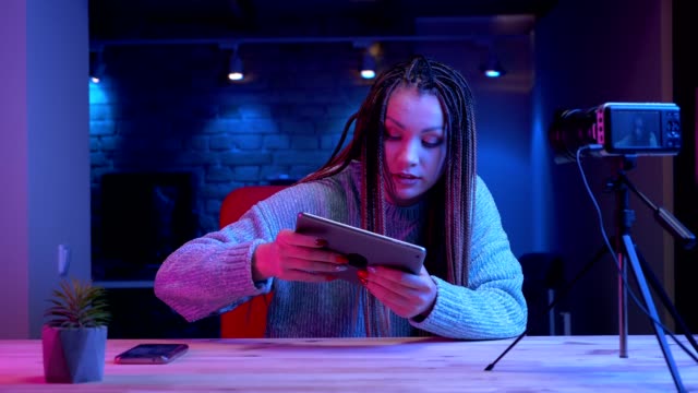 Closeup-shoot-of-young-attractive-female-blogger-with-dreadlocks-playing-video-games-on-the-tablet-live-with-the-neon-background-indoors