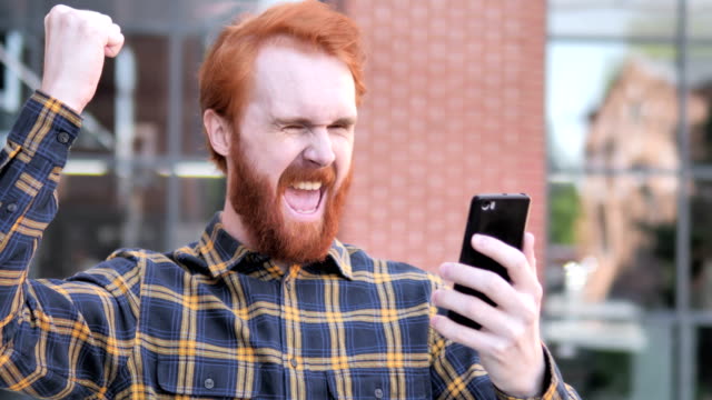 Outdoor-Redhead-Bart-junger-Mann-begeistert-für-Erfolg-auf-Smartphone