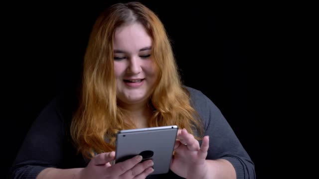 Closeup-shoot-of-young-overweight-caucasian-female-typing-on-the-tablet-and-reacting-to-social-media-posts-with-background-isolated-on-black