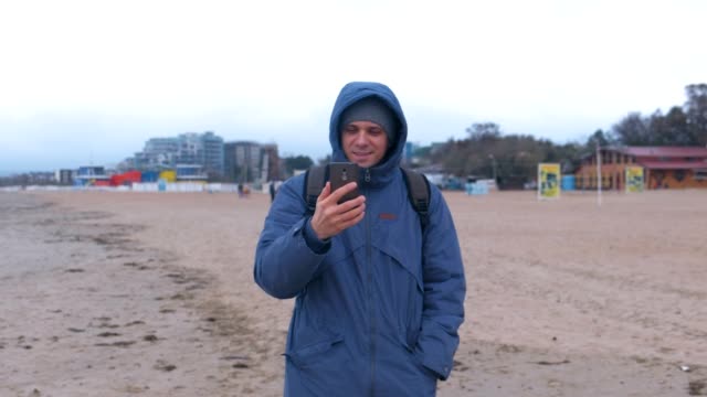 Mann-Blogger-in-einer-blauen-Daunenjacke-zu-Fuß-auf-dem-Sandstrand-am-Meer-und-sprechen-einen-Video-Chat-auf-Handy.