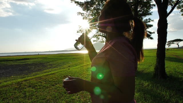 Mujer-joven-asiática-bebiendo-agua-después-de-correr-en-la-naturaleza-bajo-la-luz-del-sol-de-la-noche.-Fitness-y-concepto-de-estilo-de-vida-saludable.
