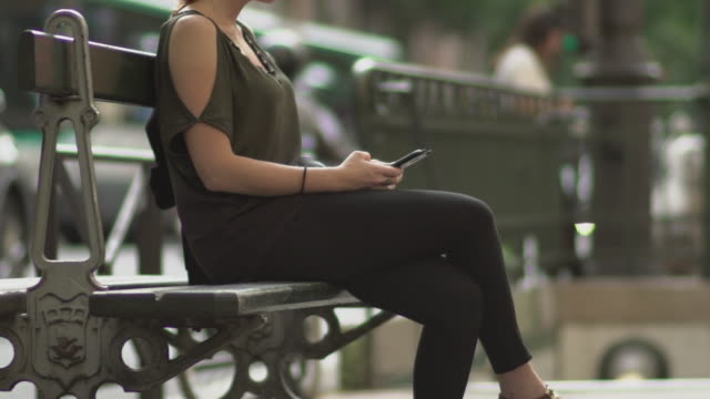 Attractive-redhead-woman-with-glasses,-freckles,-piercings-and-red-hair-writing-a-text-message-on-her-smartphone-sitting-on-street-bench,-during-sunny-summer-in-Paris.-Tilt-up-Slow-motion.-Trendy.
