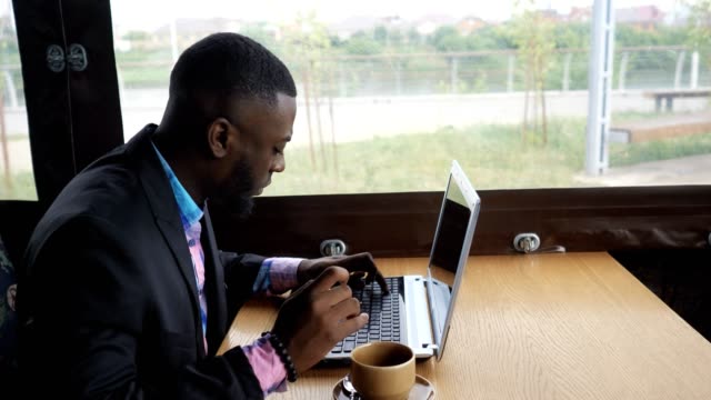 Black-businessman-is-working-typing-a-message-on-laptop-sitting-in-summer-cafe.