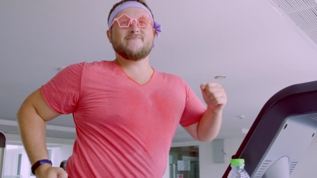 Funny-fat-male-in-pink-glasses-and-in-a-pink-t-shirt-is-engaged-on-a-treadmill-in-the-gym-depicting-a-girl.-4k
