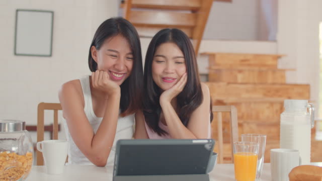 Asian-Lesbian-influencer-couple-using-tablet-record-vlog-video-on-social-media-while-having-breakfast-in-kitchen.
