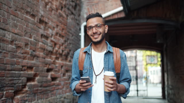 Smiling-Middle-Eastern-guy-using-smartphone-outdoors-and-holding-to-go-coffee