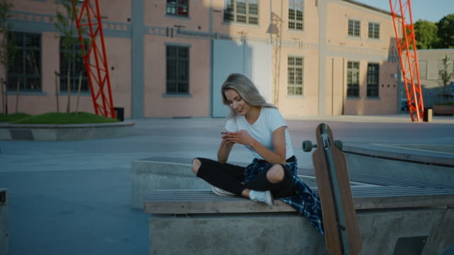 Beautiful-Blonde-Skater-Girl-Wearing-Casual-Clothes,-Uses-Smartphone,-Sharing-Stuff-on-Social-Media,-Taking-Selfies,-Messaging-Friends-and-Having-Fun-while-Sitting-on-a-Bench-in-Fashionable-Hipster