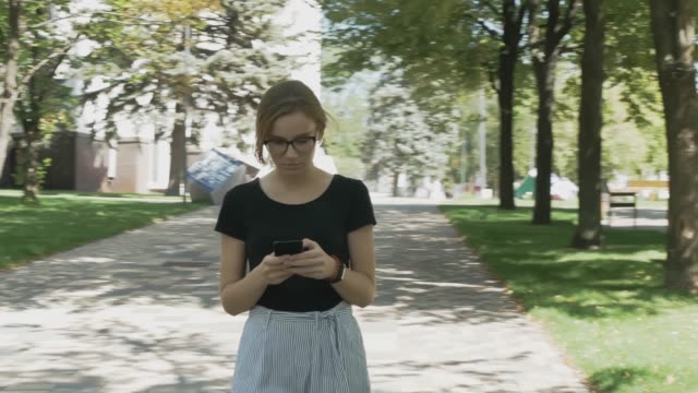 Pretty-young-caucasian-woman-in-eyeglasses-walking-towards-camera,-checking-phone-and-celebrating-good-news-in-a-park