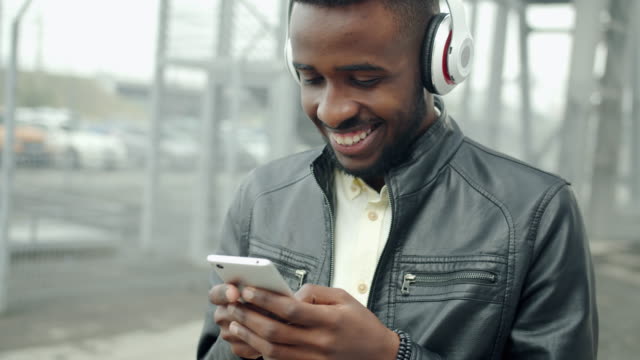 African-American-student-in-headphones-touching-smartphone-screen-outdoors