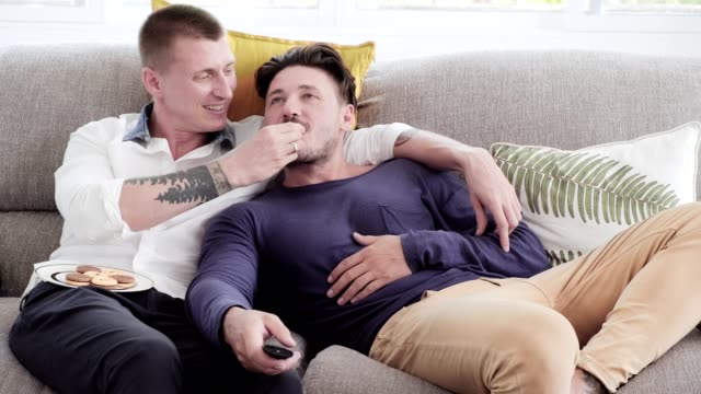 Gay-couple-relaxing-on-couch.-Watching-tv-and-feeding-food.