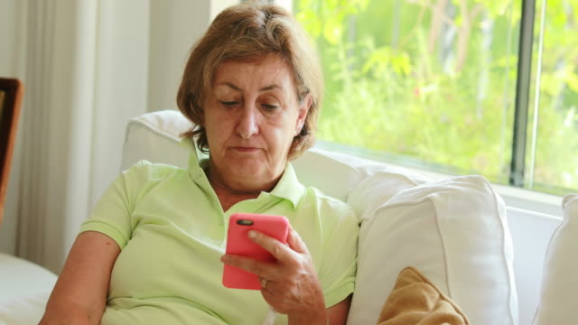 Pensada-cándida-mujer-mayor-que-sostiene-el-dispositivo-de-teléfono-inteligente-sentado-en-el-sofá-de-casa