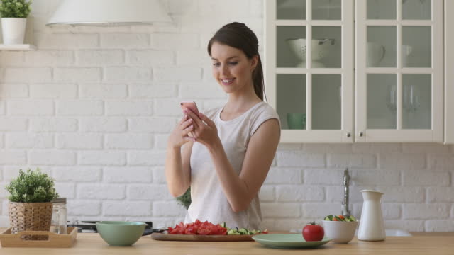 Smiling-young-woman-preparing-healthy-vegetable-salad-using-mobile-apps