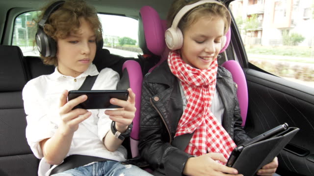 Boy-and-Girl-with-Headphones-Playing-a-Tablet-and-smartphone-in-a-Car,-Children-Using-a-Devices-in-the-auto.-Brother-and-Sister-Traveling-Together
