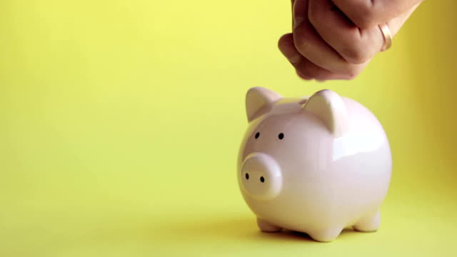 Man's-hand-putting-coins-in-piggy-bank-of-pig-shape-on-yellow-background
