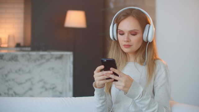 Calma-feliz-joven-mujer-en-auriculares-escalofriante-sentado-en-el-sofá-con-los-ojos-cerrados-escuchando-música-favorita-sosteniendo-el-teléfono-usando-la-aplicación-reproductor-en-línea-móvil-disfrutar-de-estado-de-ánimo-pacífico-usando-auriculare