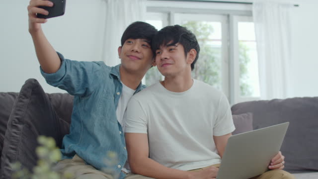 Romantic-young-gay-couple-funny-selfie-by-cellphone-at-home.-Asian-lover-male-happy-relax-fun-using-technology-mobile-phone-smiling-take-a-photo-together-while-lying-sofa-in-living-room-concept.