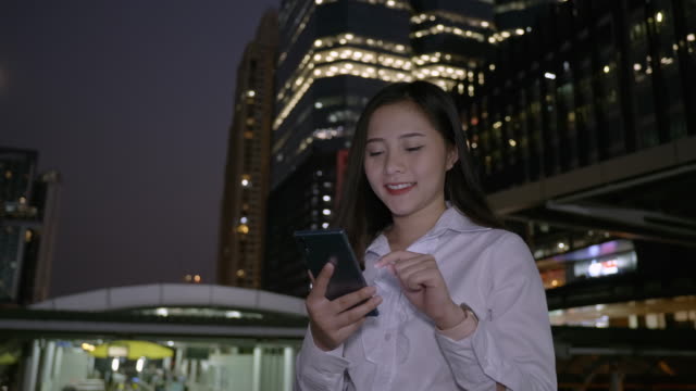 Young-smiling-business-asian-woman-using-smartphone-in-the-modern-city-center-in-the-night-of-Bangkok-Thailand.-Concept-Technology-communication-by-mobile-phone