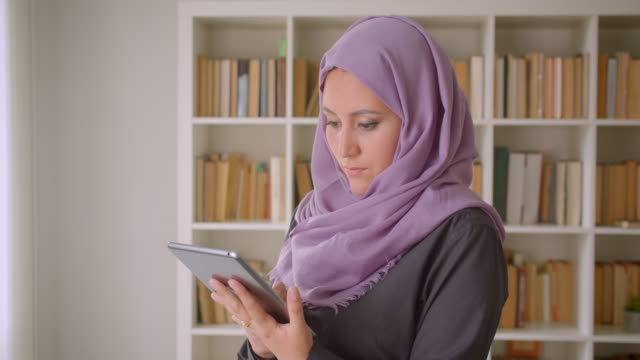 Retrato-de-primer-plano-de-joven-mujer-musulmana-en-hiyab-usando-la-tableta-y-mirando-la-cámara-de-pie-en-la-biblioteca-en-el-interior