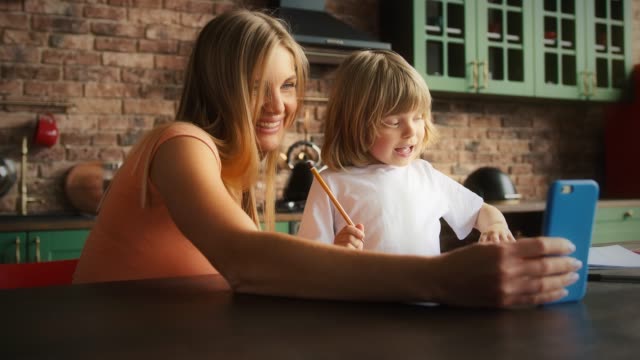 Lächelnde-Mutter-und-Kind-in-lässigem-Outfit-genießen-Online-Videoanruf-auf-dem-Handy-während-der-Hausaufgaben.-Sie-sitzen-in-der-Küche-am-Tisch.-Nahaufnahme