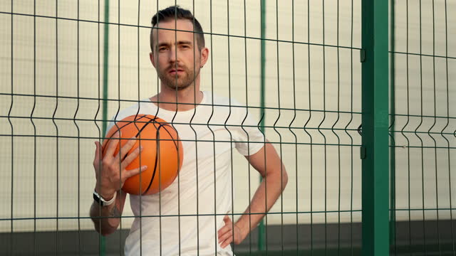 Junge-Kerl-hält-einen-Ball,-steht-hinter-einem-Zaun-von-Basketballplatz