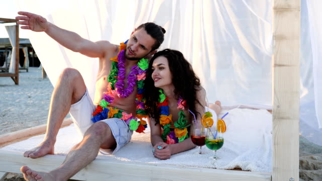 Honeymoon,-young-people-in-colorful-wreaths-sunbathe-in-bungalow-on-beach,-Backlight,-pair-of-lovers-at-Hawaii,-summer-holidays