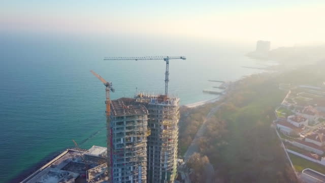 Aerial-city-view.-Construction-of-a-high-rise-skyscraper-on-the-ocean-by-two-cranes.-Around-clockwise.-The-rays-of-the-setting-sun-fall-into-the-lens