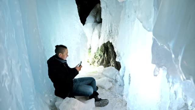 Man-communicates-in-smartphone-in-an-ice-cave.-Around-mysterious-beautiful-ice-grotto.-User-communicates-in-social-networks-in-mobile,-takes-photo-and-makes-selfie-on-phone.-He-crazy-funny-hipster.