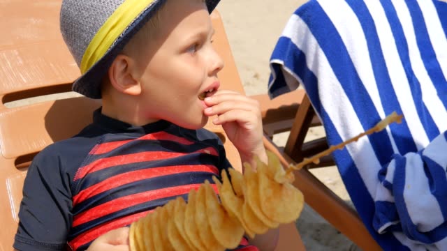 Niño-comiendo-patatas-fritas-caseras-en-un-palo,-en-la-playa-del-mar.-Pone-en-una-tumbona.