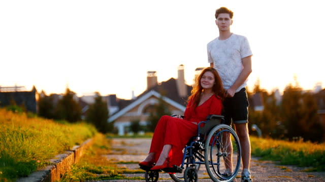 Girl-On-Wheelchair-And-Her-Boyfriend-Look-At-The-Camera-And-Smile-Slightly-In-The-Background-Of-Green