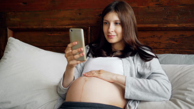 Schöne-schwangere-Mädchen-Brünette-ist-ihr-Babybauch-streicheln-und-mit-Smartphone-gemütlich-im-Bett-zu-Hause-mit-glücklichen-Lächeln.-Schwangerschaft,-Familie-und-Technologie-Konzept.