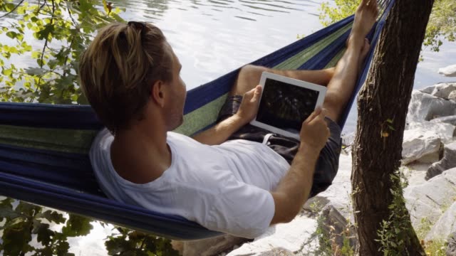 Hombre-joven-con-tableta-digital-en-hamaca,-mintiendo-entre-los-árboles-por-el-río-en-verano
