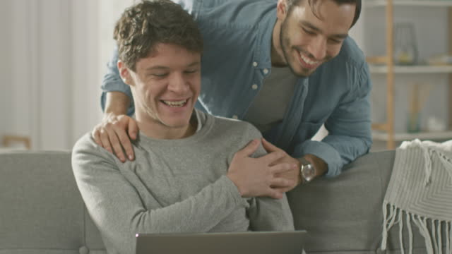 Dulce-pareja-gay-hombre-pasa-tiempo-en-casa.-Joven-usa-un-ordenador-portátil,-su-compañero-viene-desde-atrás-y-suavemente-le-abraza.-Se-ríen-y-tocar-manos.-Habitación-tiene-un-Interior-moderno.