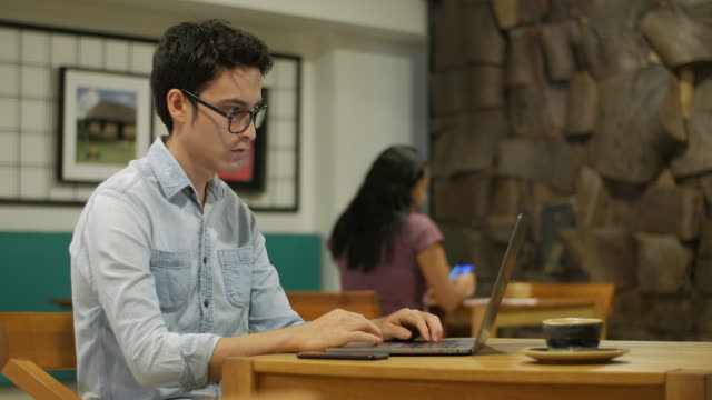 Hombre-hispano-sentado-en-la-cafetería-escribiendo-en-un-ordenador-portátil,-chica-en-el-fondo