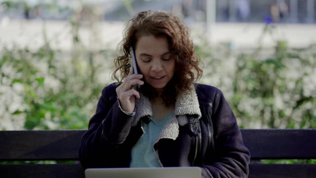 Smiling-curly-woman-talking-on-smartphone-while-sitting-on-bench.