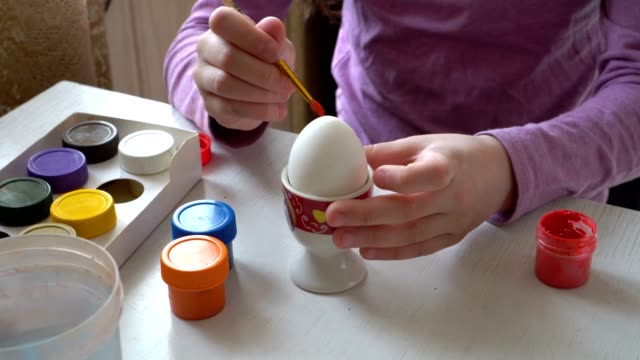 Ein-kleines-Mädchen-zieht-mit-einem-Pinsel-und-farbigen-Farben-auf-ein-weißes-Ei,-sitzt-zu-Hause-am-Tisch.-Close-up.-4K.