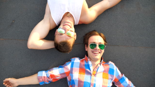 Top-view-of-handsome-gay-boys-in-sunglasses-lying-on-rooftop-of-high-rise-building-and-laughing.-Young-happy-male-couple-looking-at-camera-and-showing-different-grimases-on-roof.-Close-up-Slow-motion