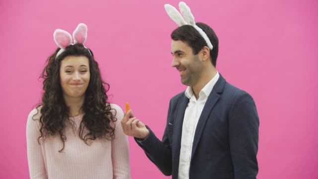 Young-couple-are-beautiful-on-pink-background.-During-this-time-they-are-dressed-in-rabble-ears.-Looking-at-each-other.-The-man-shows-the-carrots-and,-the-wife-begins-to-laugh-sincerely.-A-young-man-disagreed-and-threw-carrots-into-his-wife.