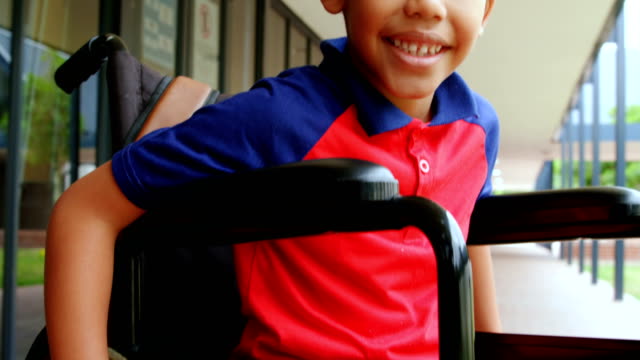 Vista-frontal-de-feliz-colegial-afroamericano-con-discapacidades-sentado-en-silla-de-ruedas-en-el-corredor-escolar-4k