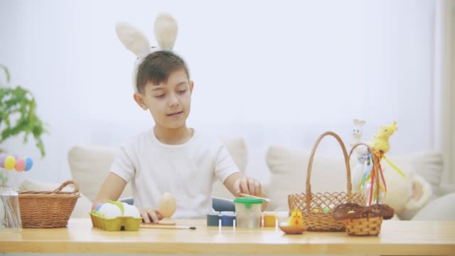 Cute-little-boy-with-bunny-ears-is-sitting-at-the-table-full-of-Easter-decorations-and-raising-his-thumb-fingers-up,-smiling.-Pretty-Easter-boy-is-taking-a-nude-brush-and-starting-to-paint-an-Easter-egg.