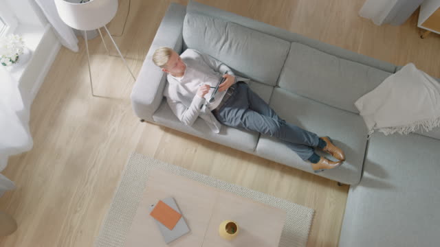 Young-Man-is-Lying-on-a-Sofa,-Working-or-Sketcing-on-a-Tablet-Computer.-Cozy-Living-Room-with-Modern-Interior,-Grey-Sofa-and-Wooden-Flooring.-Top-View-Camera-Footage.